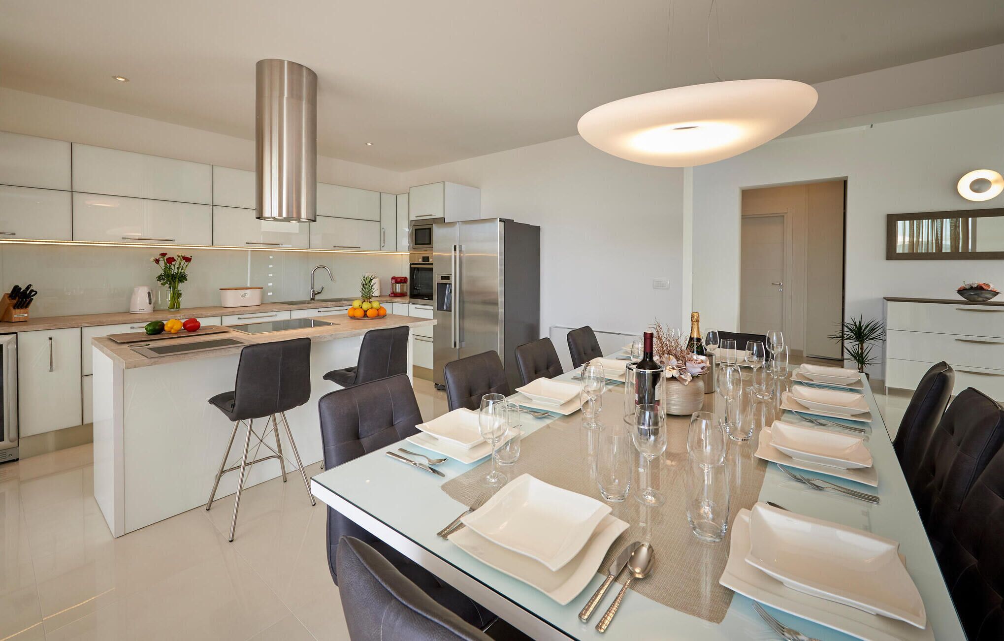 Une cuisine moderne dotée d’un îlot central, d’appareils électroménagers en acier inoxydable et d’un coin repas aménagé pour six personnes, avec une table et des chaises en verre.