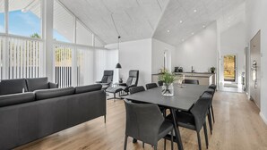 Ampla sala de estar com um sofá grande, mesa de jantar e cadeiras, cozinha com pia e bancada, e grandes janelas que permitem entrada de luz natural.