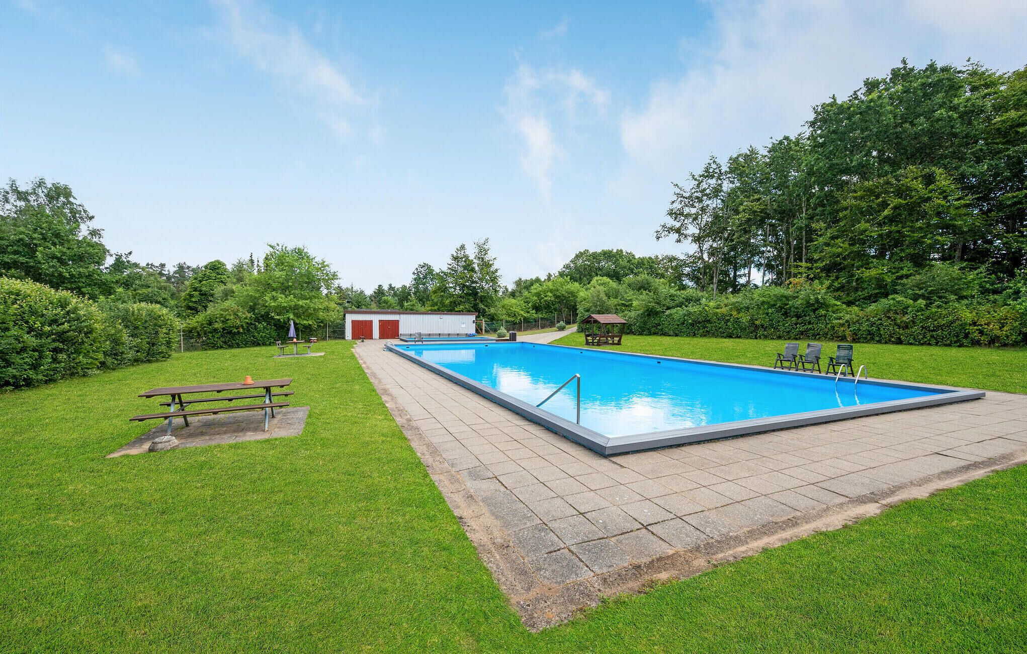 A rectangular swimming pool with a tiled deck, surrounded by a grassy area with a picnic table and a gazebo.