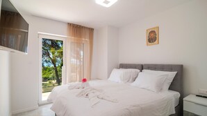 A bedroom with a bed, a nightstand, a wall-mounted TV, and a window with a view of trees. - Cozy home in Sumartin (Sumartin)