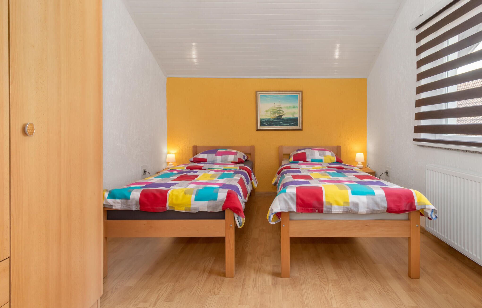 Une chambre avec deux lits en bois, chacun agrémenté d’une literie aux couleurs vives, un mur jaune, un tableau au-dessus des lits et une fenêtre avec des stores.