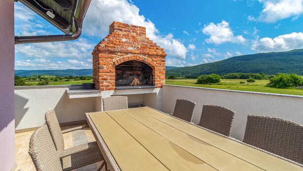 An outdoor patio with a brick-built barbecue grill, wicker furniture, and a view of the countryside.