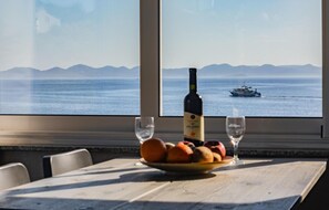 Taula amb ampolla de vi, dues flautes i un plat de fruita. Es veu el mar i un vaixell a través d'una finestra.