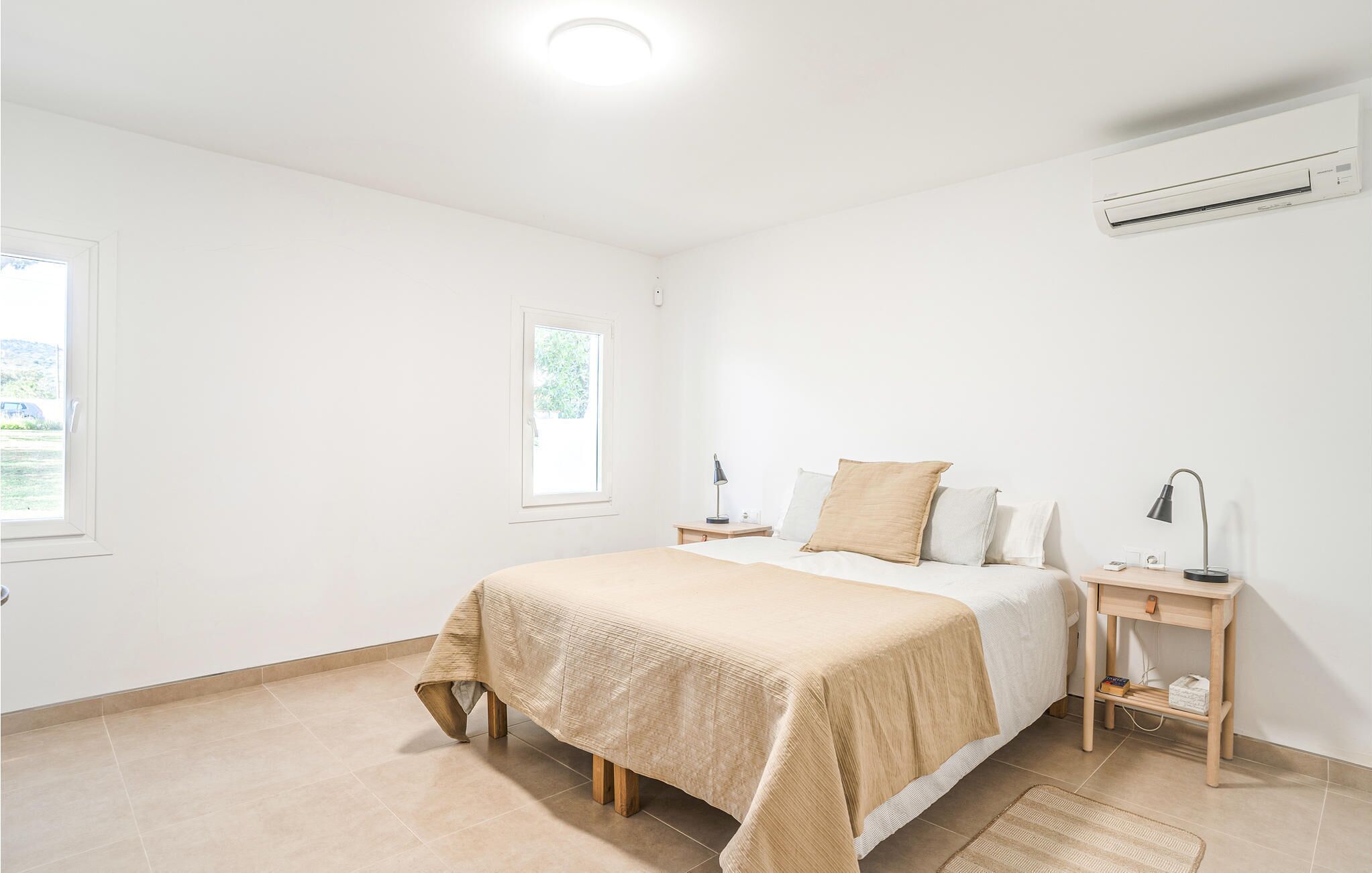 A minimalist bedroom with a bed, bedside tables, a lamp, and an air conditioning unit.