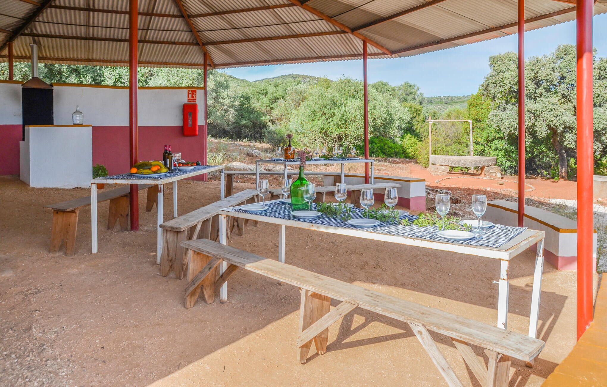 An outdoor dining area with tables and benches, surrounded by a covered structure and a view of a hilly landscape.
