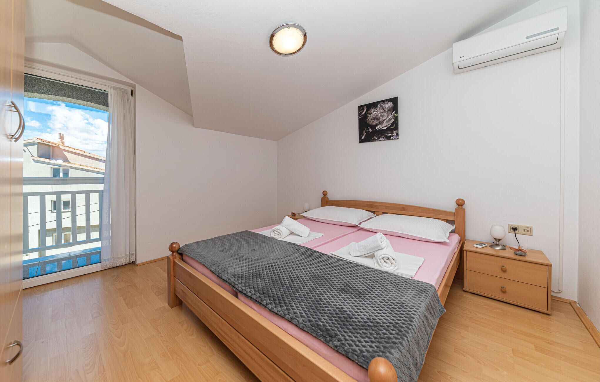 Een slaapkamer met een houten bed, een nachtkastje, een airconditioner en een raam met uitzicht op gebouwen.