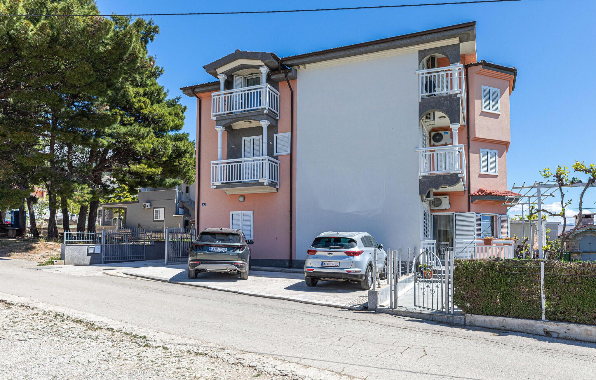 A multi-story residential building with balconies, a gated driveway, and parked cars.