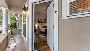 Un corridoio stretto con una porta che dà su una camera da letto. La stanza presenta un letto, una sedia e un comodino. A destra della porta, si apre una finestra con persiane.