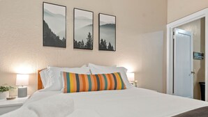 Un letto rifatto con lenzuola bianche e un cuscino a righe colorate, una lampada da comodino e due quadri appesi alla parete.