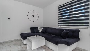 Un salon moderne avec un canapé d’angle, une petite table d’appoint blanche, une horloge murale et une fenêtre avec des stores.