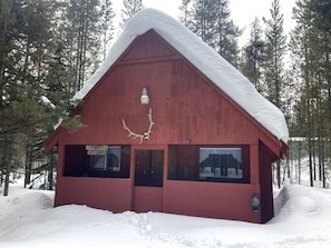 Une cabane en bois rouge, avec un toit recouvert de neige, entourée d’arbres.