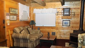 A cozy cabin interior with a sofa, a small table, a lamp, and a wood-burning stove.