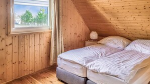 Un dormitorio con techo inclinado de madera, una cama con ropa blanca, una mesita de noche con lámpara y una ventana con vistas a árboles y un edificio.