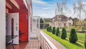 Una terraza de madera con baranda blanca, un edificio rojo y un césped cuidado con arbustos podados y una casa al fondo.