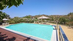 A clear blue swimming pool with a wooden deck, surrounded by white chairs and umbrellas, set against a backdrop of hills and clear skies. - 6 bedroom beautiful home in Perdifumo (Perdifumo)
