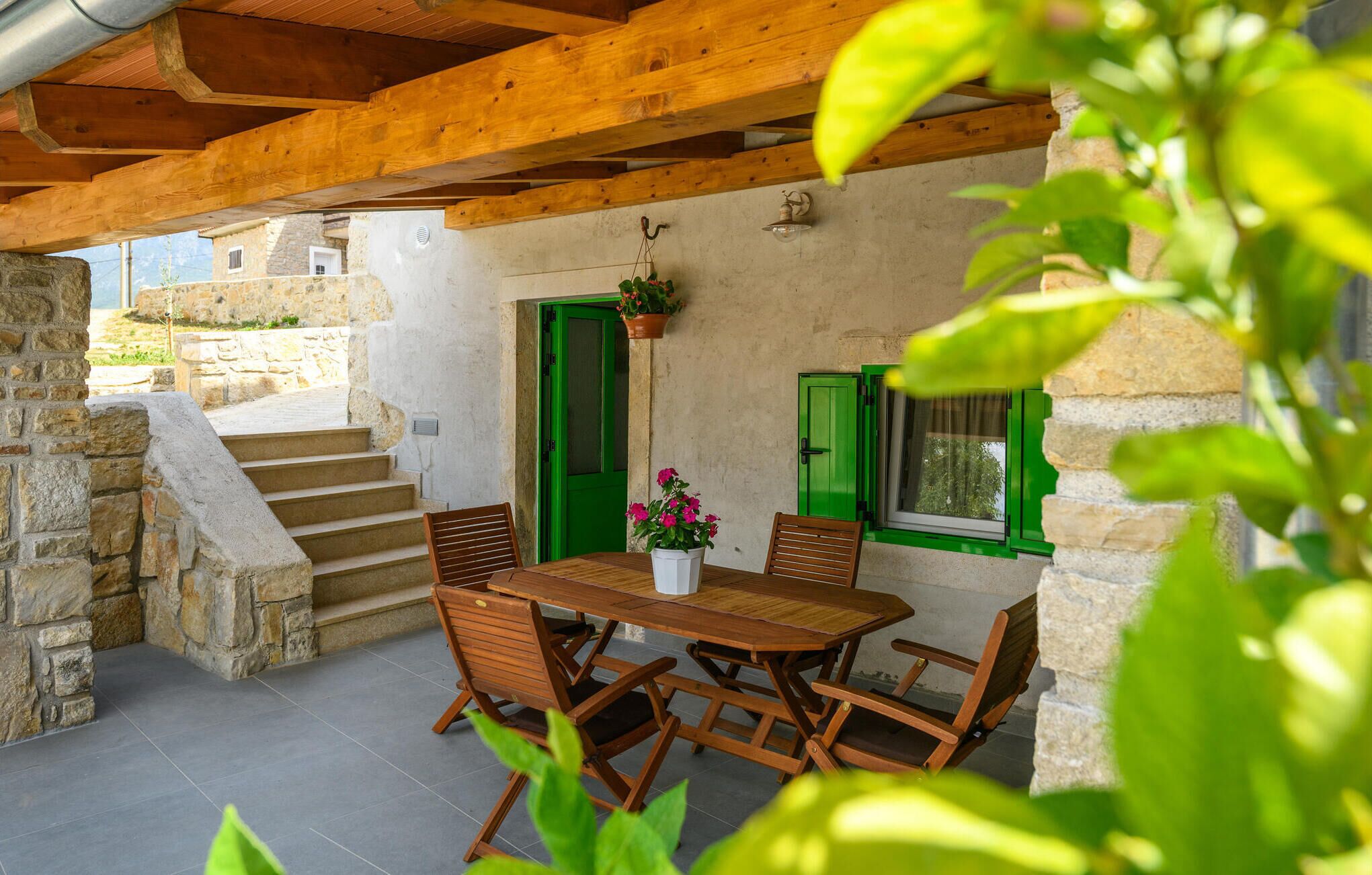 A wooden table with chairs and a potted plant in a stone-walled outdoor area with green shutters and a staircase.