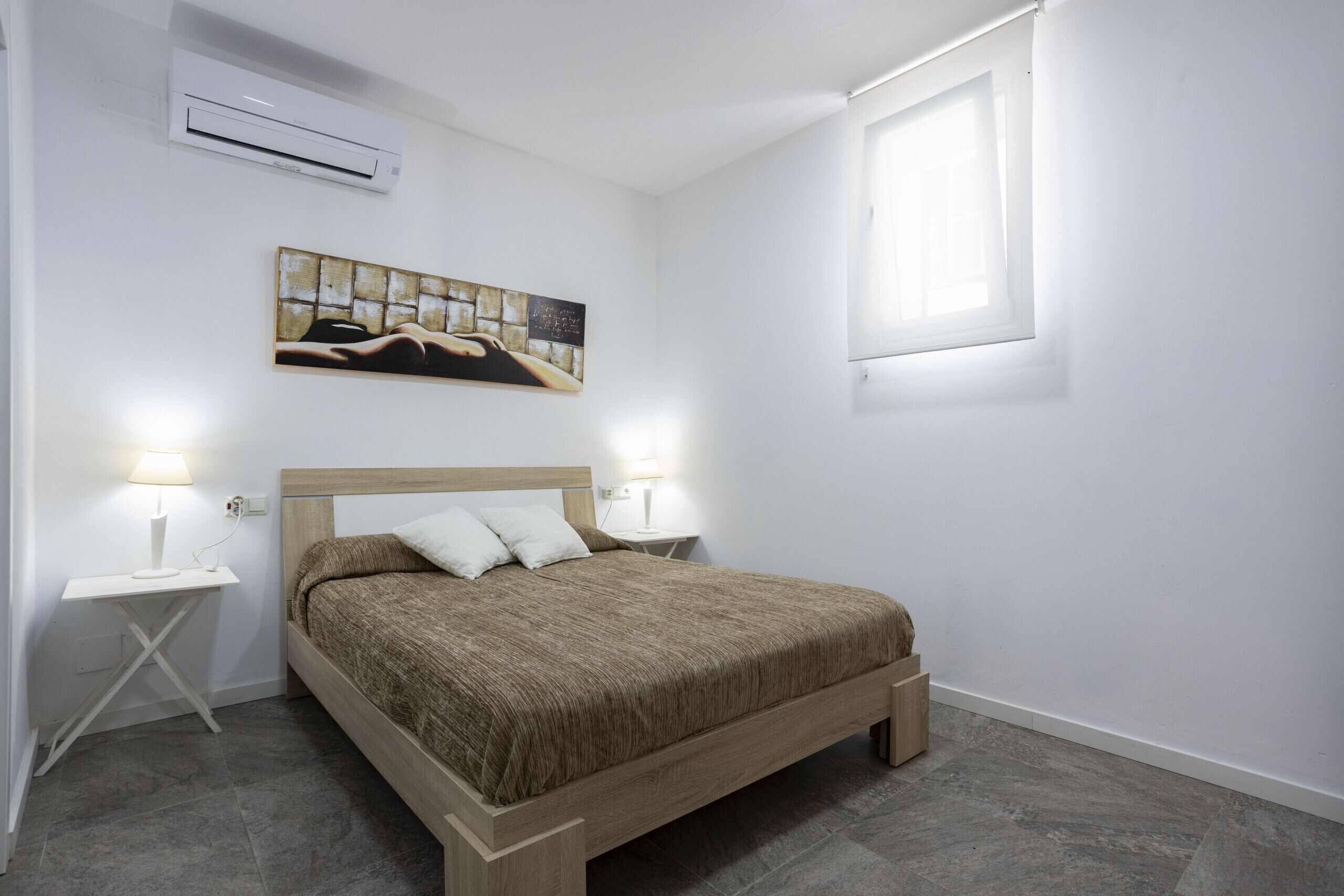 A bedroom with a wooden bed, white walls, a painting above the bed, and a window on the right.