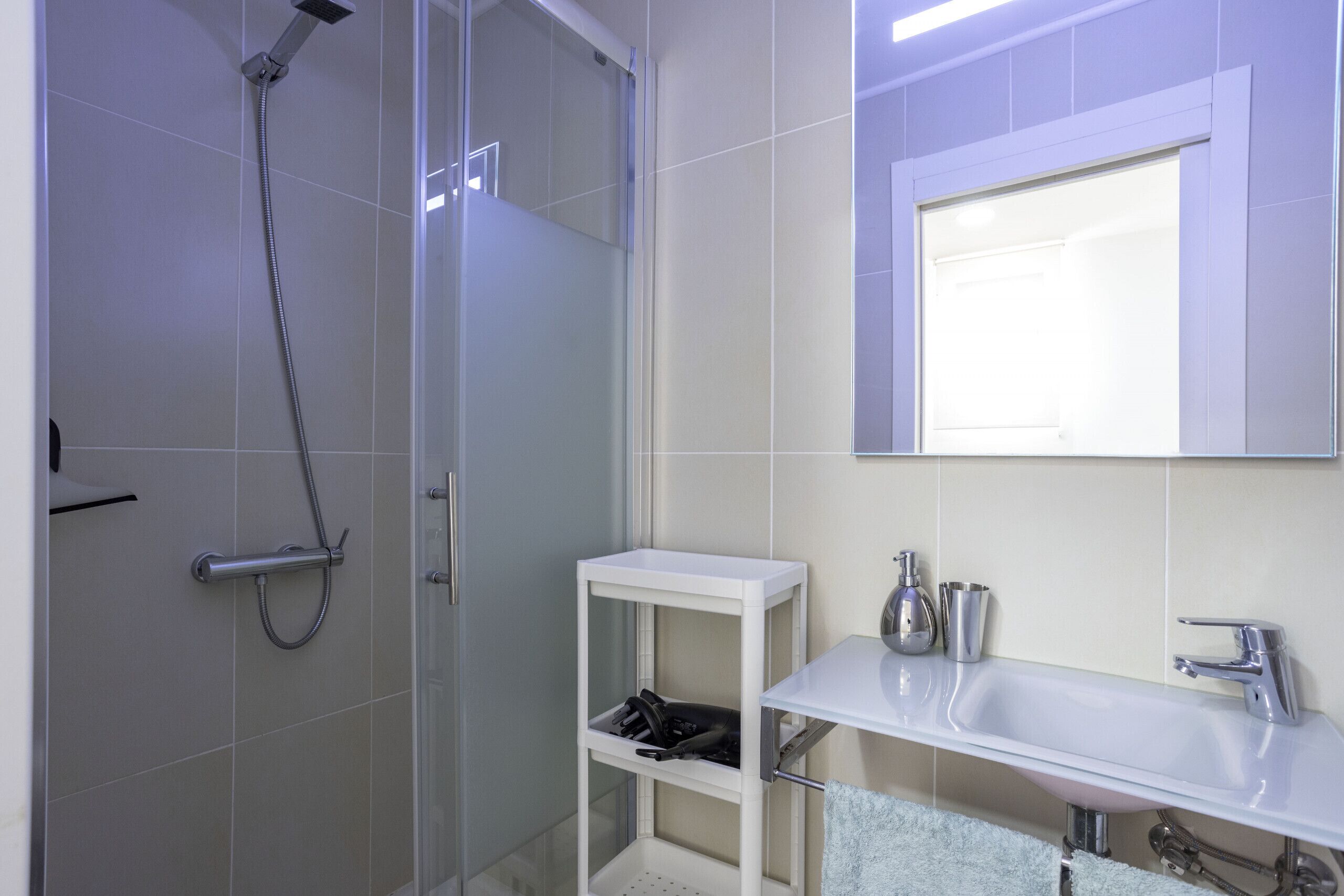 Un baño moderno con ducha envuelta en vidrio, un lavamanos blanco con grifería cromada y un estante blanco pequeño con artículos de aseo.