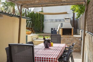 Un patio al aire libre con una mesa lista para comer, rodeada de sillas y una parrilla de barbacoa al fondo.