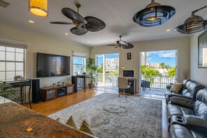A spacious living room with a large flat-screen TV, ceiling fans, a leather sofa, and a view of a balcony with outdoor plants.