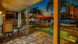 An outdoor patio area with a pool, seating, and a view of palm trees and a sunset.