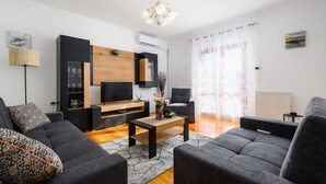 Een moderne woonkamer met een flatscreen televisie, grijze banken, een houten salontafel en een kroonluchter.