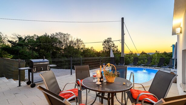 Een terras buiten met een zwembad, lounge stoelen, een barbecue en een tafel gedekt voor een maaltijd.