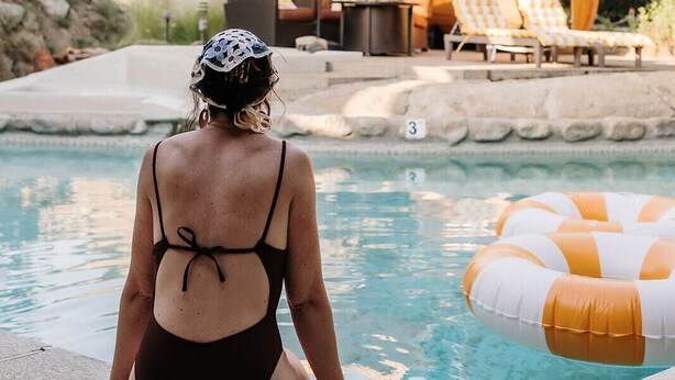 Una mujer sentada junto a una piscina, con sillones de descanso y una sombrilla al fondo.