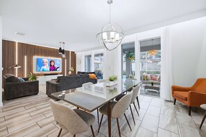 A modern living room with a flat-screen TV, a dining area with a glass table, and a large window with a view.