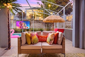 A cozy outdoor seating area with a wicker sofa, colorful cushions, a thatched umbrella, and a TV screen displaying a cartoon. - Walk2Clubhouse|Pool Movies|WaterPark|Arcade|Themed (Davenport)