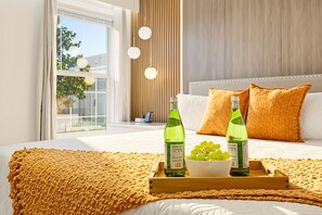 A bedroom with a bed, pillows, a tray with bottles and grapes, and a window with a view.
