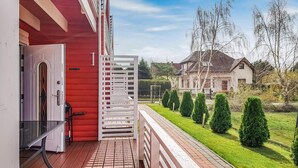 A wooden deck with a white railing, a red building, and a well-maintained lawn with trimmed bushes and a house in the background.