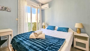 Um quarto com cama, duas mesas de cabeceira, escrivaninha, janela com cortinas e ar condicionado.