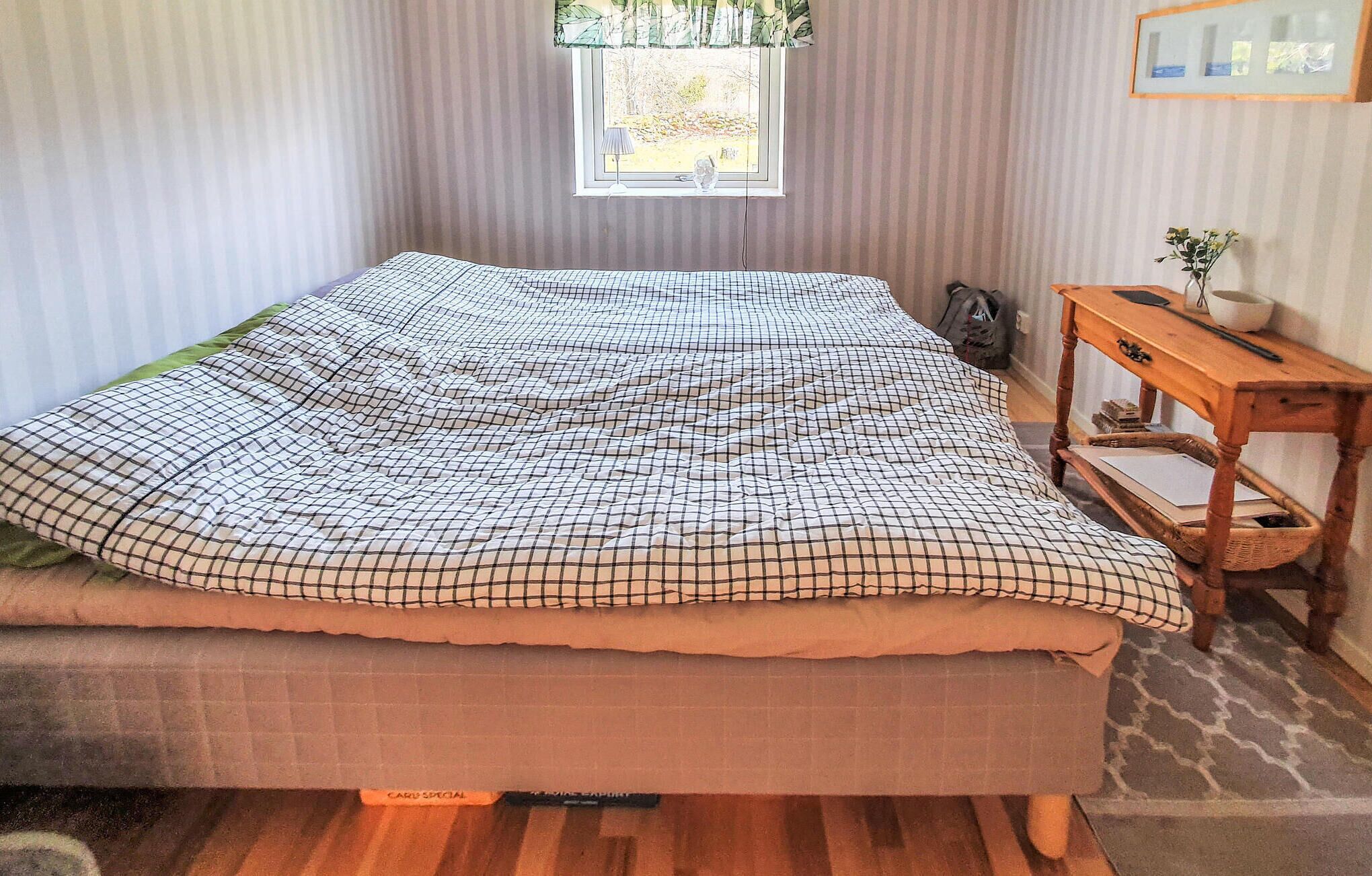 A bed with a checkered bedspread, a window with a view of trees, a small table with a plant, and a wicker basket.