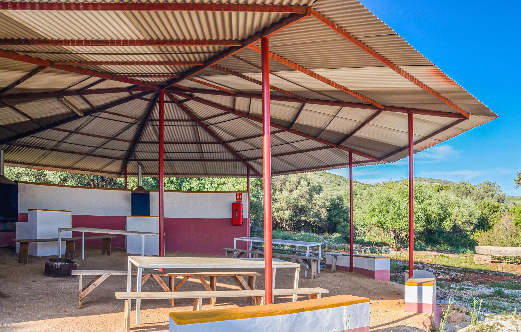 A covered outdoor area with picnic tables and benches, surrounded by greenery.