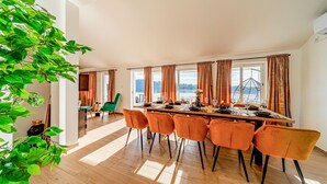 A dining area with a long table, orange chairs, and large windows offering a view of the outdoors.