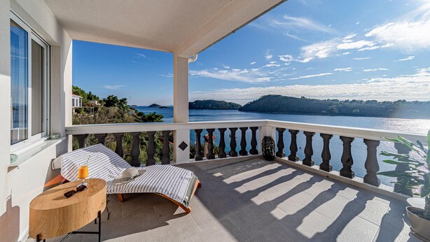 A balcony with a view of a lake and hills, a wooden table with a drink, and striped cushions.