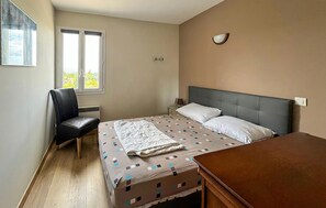 Un dormitorio con cama, una silla, una mesita de noche, una lámpara y ventana con vistas.