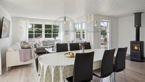 Ein modernes Wohnzimmer mit Essbereich, weißen Wänden, großen Fenstern und einem Kaminofen.