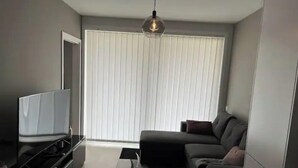 Ein modernes Wohnzimmer mit einem Flachbildfernseher, einem dunklen Sofa und einem großen Fenster mit Jalousien.