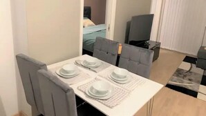 Ein Essbereich mit einem weißen Tisch, gedeckt für vier Personen, grauen Polsterstühlen und Blick in ein Schlafzimmer mit Bett und Fernseher.