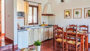 A kitchen with a dining area, featuring a tiled floor, wooden chairs, a dining table set for four, a microwave, and a window with curtains.