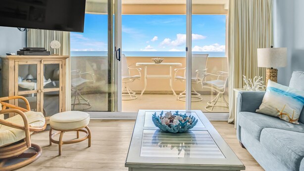 A living room with a television, a glass coffee table, wicker chairs, a sofa, and a view of the ocean through a sliding glass door. - Charming Oceanfront Condo, 2 Open private balconies (Ocean City)
