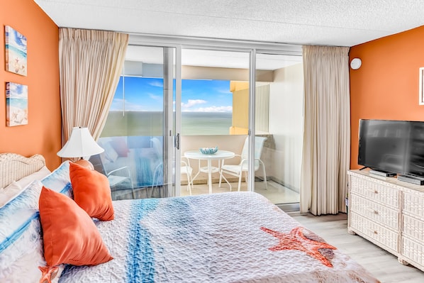 A hotel room with a bed, a TV, a balcony with a view of the sea, and a dining area.