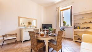 Een eetruimte met een ronde tafel, rieten stoelen, een witte kast, een televisie en een raam met uitzicht op bomen.