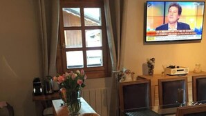 Ein Essbereich mit einem Holztisch und Stühlen, einer Vase mit Blumen sowie einem Fernseher, der eine Nachrichtensendung zeigt.