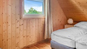 Et soveværelse med trævægge, et vindue med udsigt til grøn vegetation, en seng med hvide sengetøj og et natbord med en lampe.