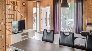 Et træinteriør med et fjernsyn på et hvidt skærmstativ, et spisebord med stole og udsigt til det fri via store vinduer.