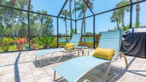 A sunlit outdoor lounge area with two lounge chairs, one with a yellow pillow, and a view of palm trees and a body of water through a glass enclosure. - Nature's Splendor! Lovely Setting. (Rotonda West)