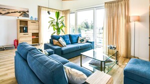 Een moderne woonkamer met een blauwe bank, een glazen salontafel en een groot raam met gordijnen.